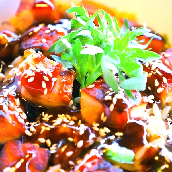 Close-up of glazed chicken or pork pieces with sesame seeds and fresh green herbs.